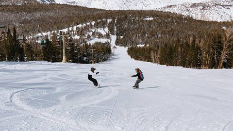 Two snowboarders descending a wide, groomed beginner run at June Mountain